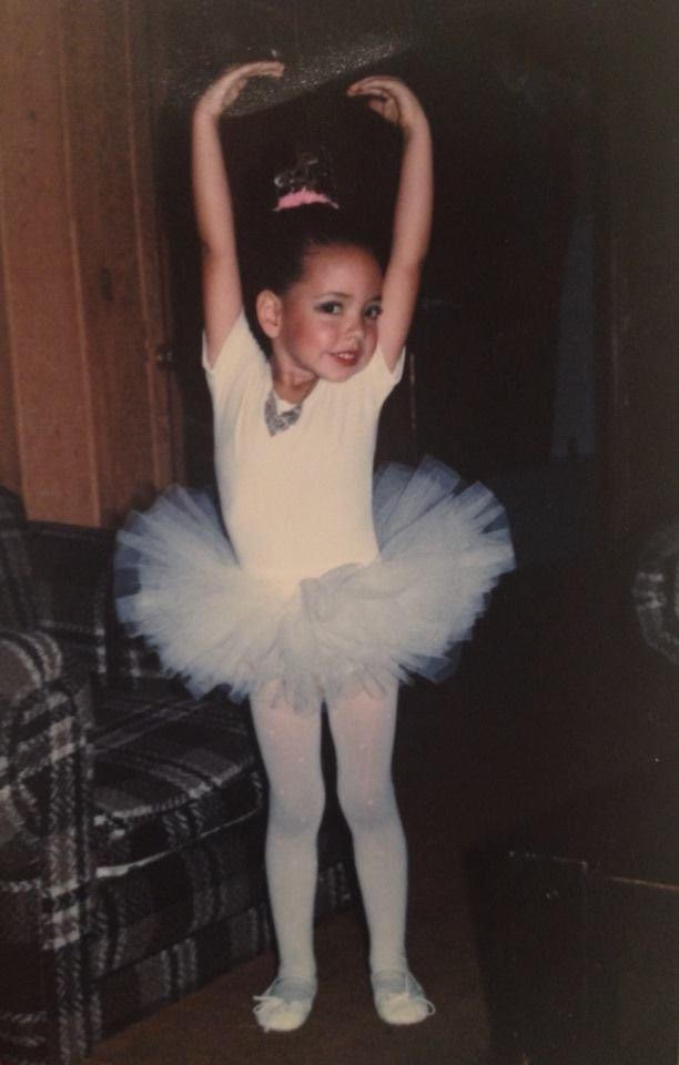 Pamela as a young girl in a white leotard and tutu, arms raised over her head in a ballet pose